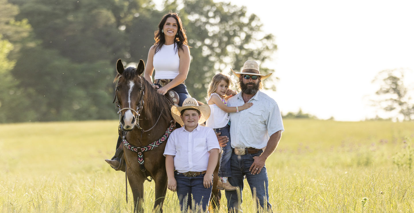 The Smith Family, Kip, Kalie, Tarver, and Cecillia, dressed in Cowboy attire, in a field, while Kalie with black hair is riding a brown horse. 